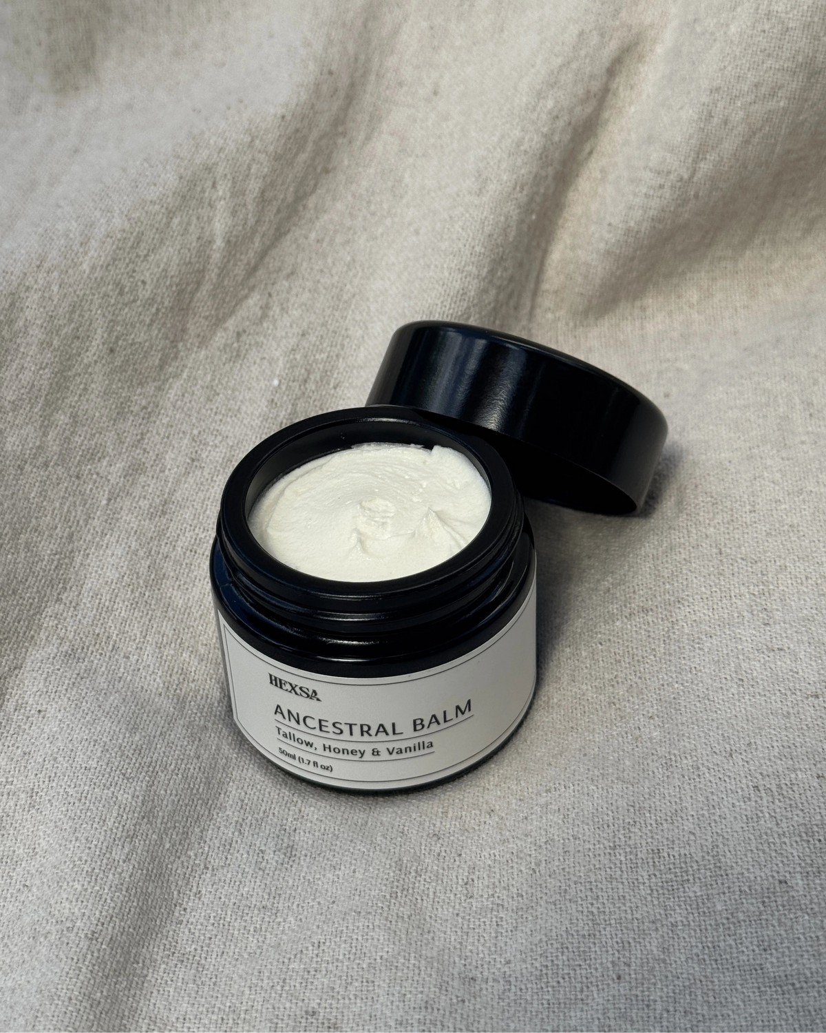 Open jar of 'Ancestral Balm' on a textured beige fabric background
